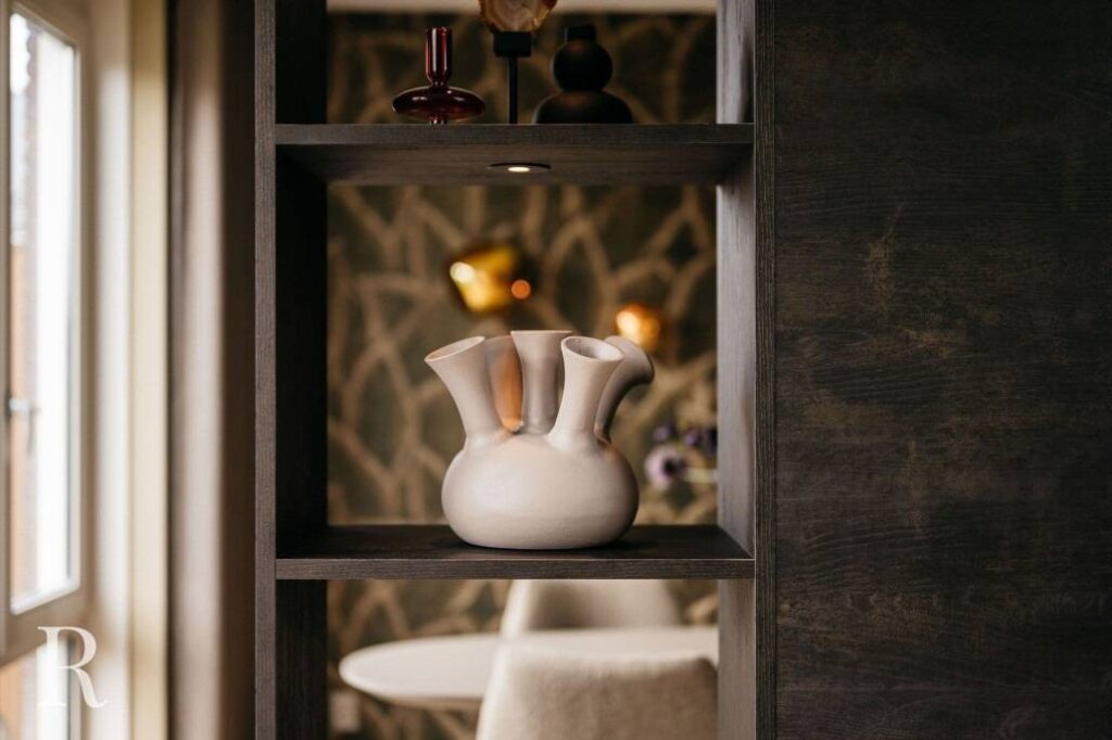 Een witte keramische vaas met meerdere openingen staat op een donkerhouten cinewall-plank, tegen een zacht verlicht, stijlvol interieur en een geometrische stenen muur op de achtergrond.