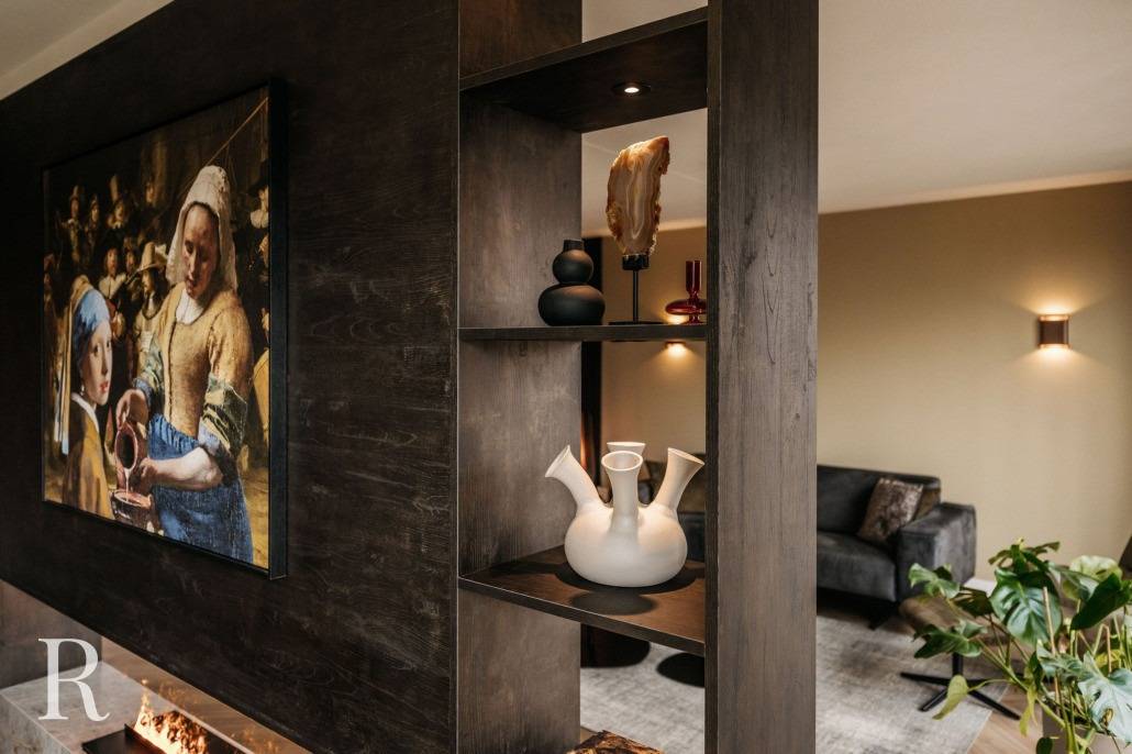 Een moderne woonkamer met een donkerhouten cinewall scheidingswand met planken versierd met decoratieve vazen en kunstwerken. Aan de muur hangt een groot ingelijst schilderij van mensen in historische kleding; een bank en weelderige planten maken de verfijnde ruimte compleet.