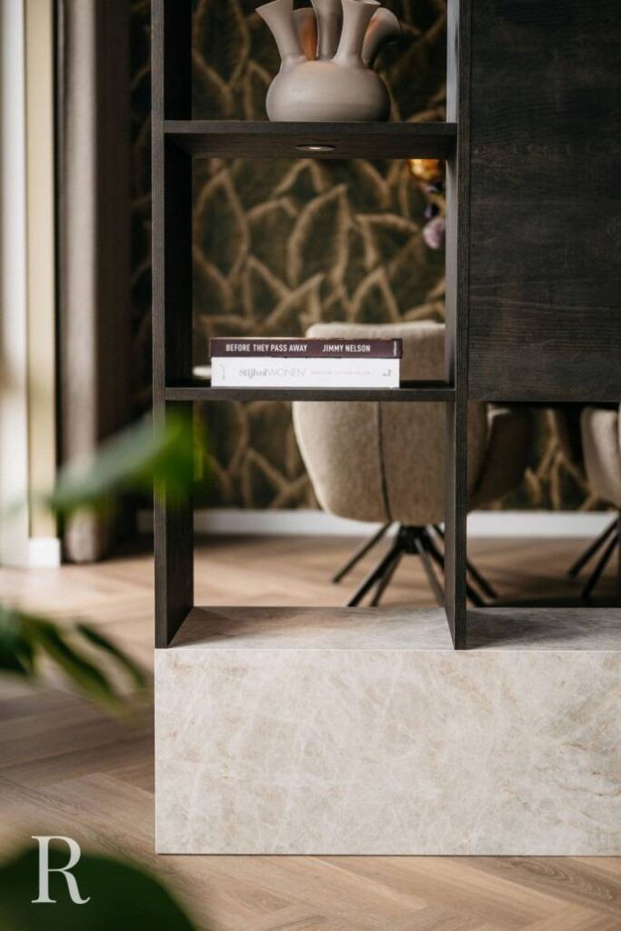 Een close-up van een modern schapmeubel, vakkundig ingebouwd in een donkerhouten cinewall, met twee vazen op de bovenste plank en boeken eronder, geaccentueerd door neutraal getinte fauteuils en behang met patroon op de achtergrond.