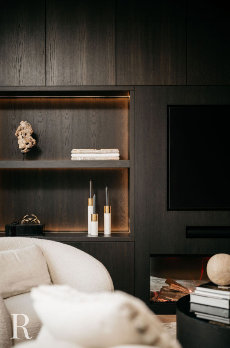 Moderne woonkamer met donkerhouten ingebouwde planken met decoratieve voorwerpen, waaronder boeken, een beeld en kaarsen. Een tv en open haard zijn in de muur ingebouwd. Een fauteuil met lichte textuur staat op de voorgrond.