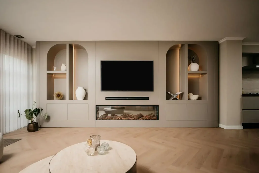 Een moderne woonkamer heeft een ingebouwde entertainmentunit met een grote tv, decoratieve planken met vazen en beeldhouwwerken, een elektrische open haard, lichthouten vloeren en een ronde salontafel met glazen decor.
