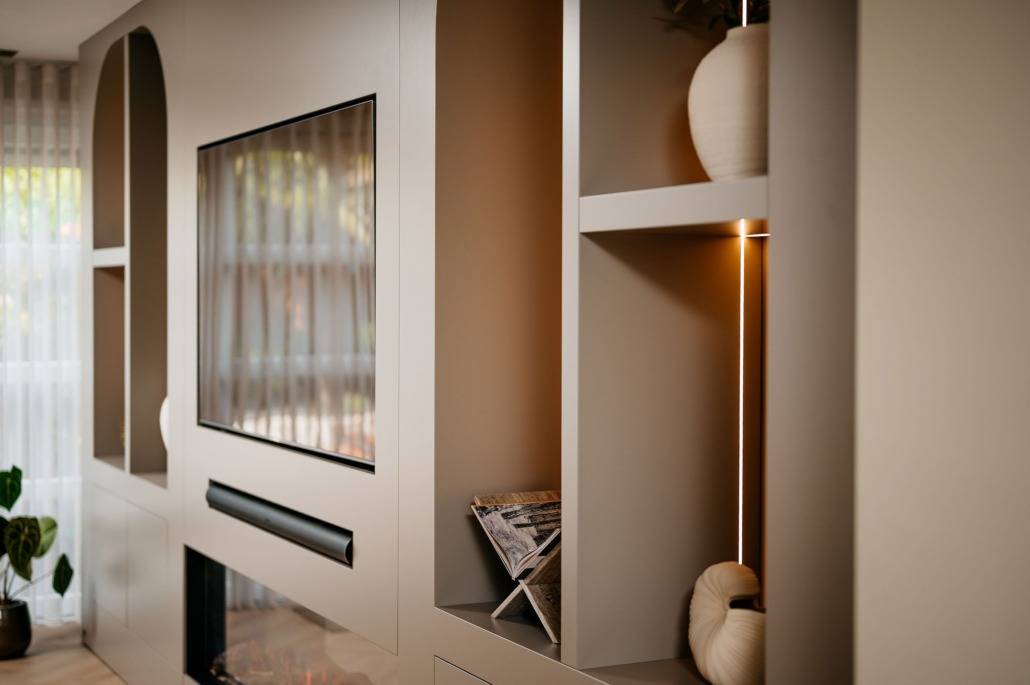 Modern wandmeubel in de woonkamer met ingebouwde planken met decoratieve vazen en boeken, een flatscreentelevisie en een open haard eronder. Zachte verlichting benadrukt de beige tinten en de minimalistische inrichting.