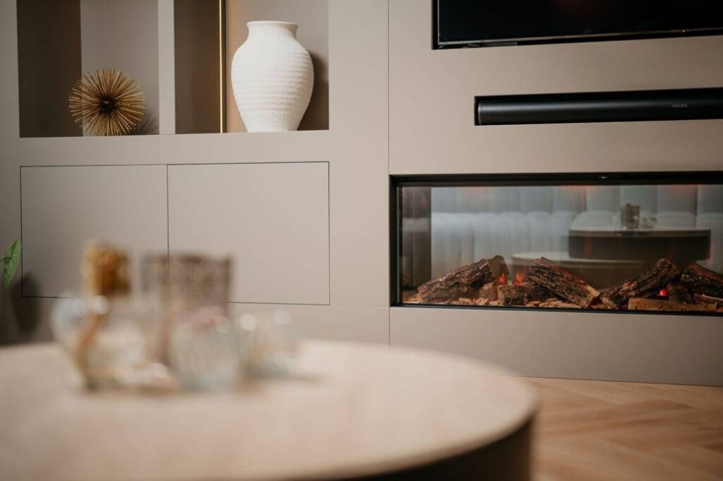 Een moderne woonkamer met een lichtgekleurde ingebouwde plank, een witte keramieken vaas, een decoratieve stekelige sculptuur, een met glas bedekte elektrische open haard en een ronde houten salontafel met glaswerk op de voorgrond.