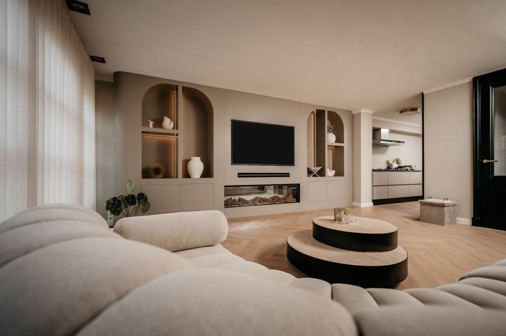 Moderne woonkamer met een gebogen beige bank, ronde salontafels, ingebouwde planken met decoratieve vazen, een tv aan de muur en een open keuken op de achtergrond. Lichte houten vloeren en transparante gordijnen voegen warmte toe.