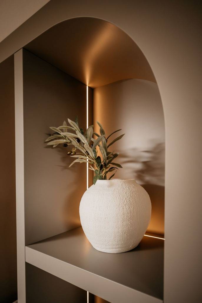 Een vaas van wit keramiek met groene bladeren staat op een plank in een moderne, zacht verlichte alkoof met gebogen bogen en warme beige tinten.