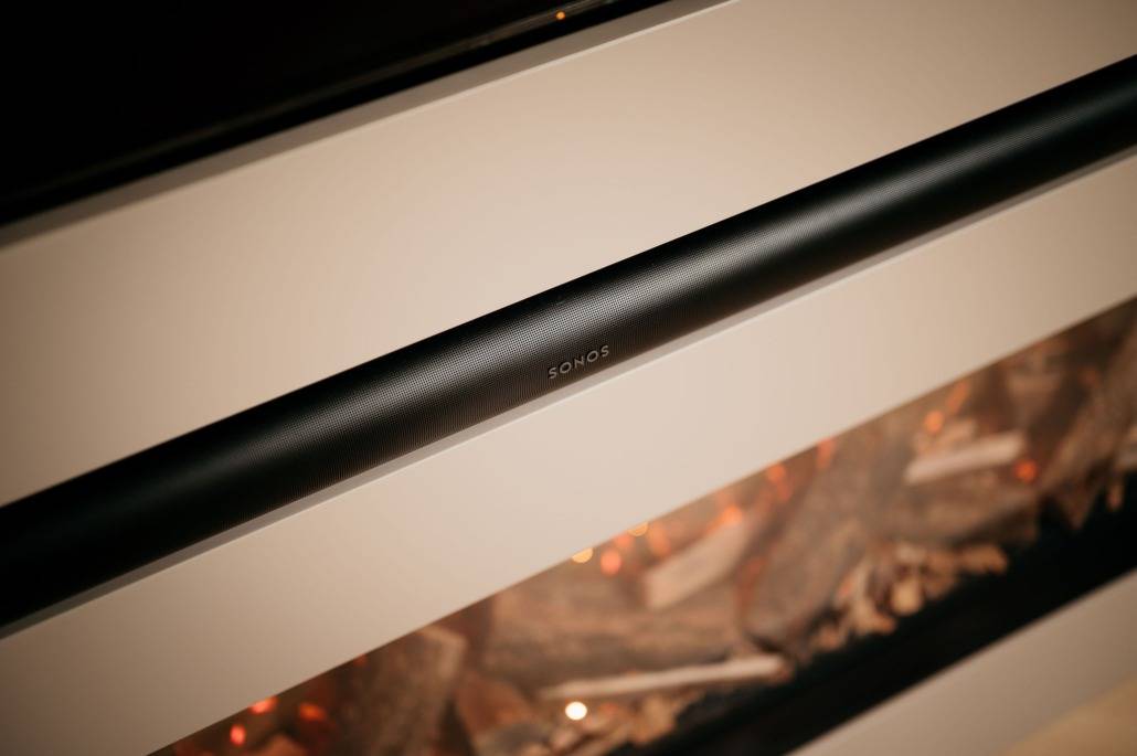 Een close-up van een zwarte Sonos soundbar gemonteerd boven een moderne elektrische open haard met houtdecoraties en warme gloeilampen.
