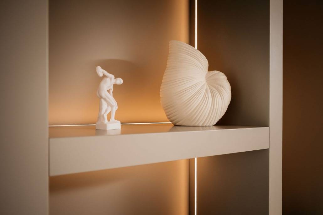 Op een witte decoratieve plank staan een klein beeldje van een man met een schijf en een groot, wit, schelpvormig beeld, beide verlicht door een warm, zacht tegenlicht.