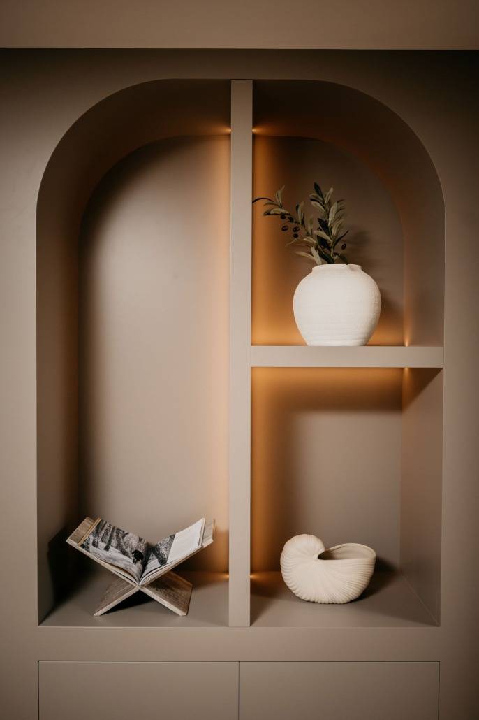 Een ingebouwde, gewelfde nis met zachte verlichting toont een witte vaas met olijftakken, een sculpturaal keramisch stuk en een open tijdschrift op een standaard tegen een neutrale beige achtergrond.