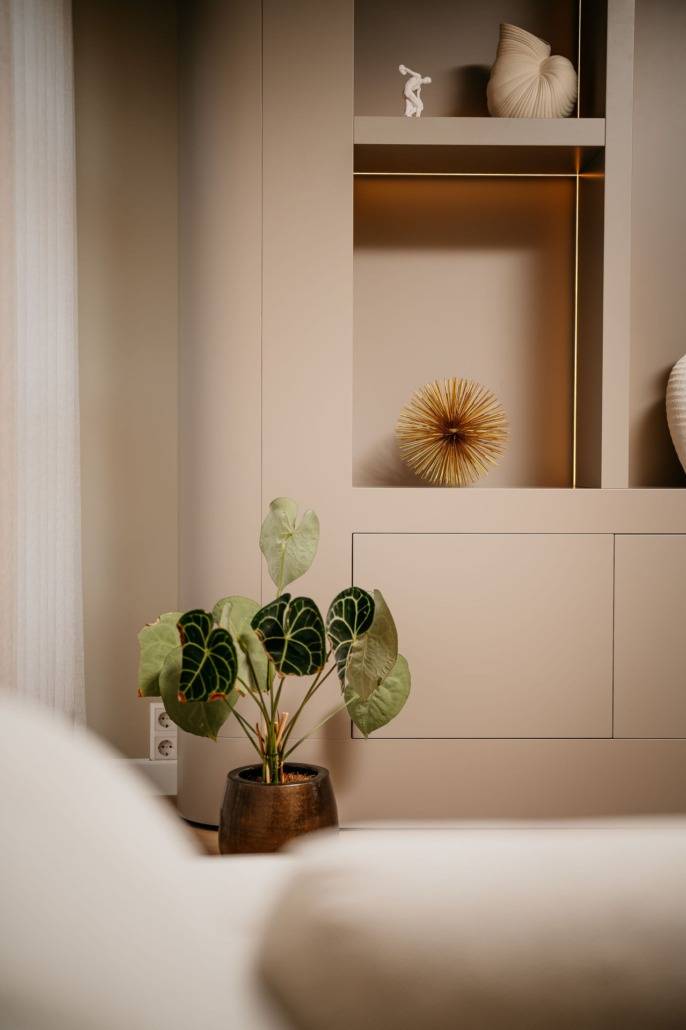 Een moderne, minimalistische woonruimte met beige muren en planken met decoratieve voorwerpen. Een potplant met grote groene bladeren staat op de voorgrond bij een zachte, lichtgekleurde bank.