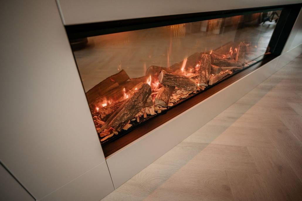 Een moderne ingebouwde elektrische open haard met realistisch gloeiende sintels en nephoutblokken, geplaatst in een lichtgekleurde muur naast een lichte houten vloer.