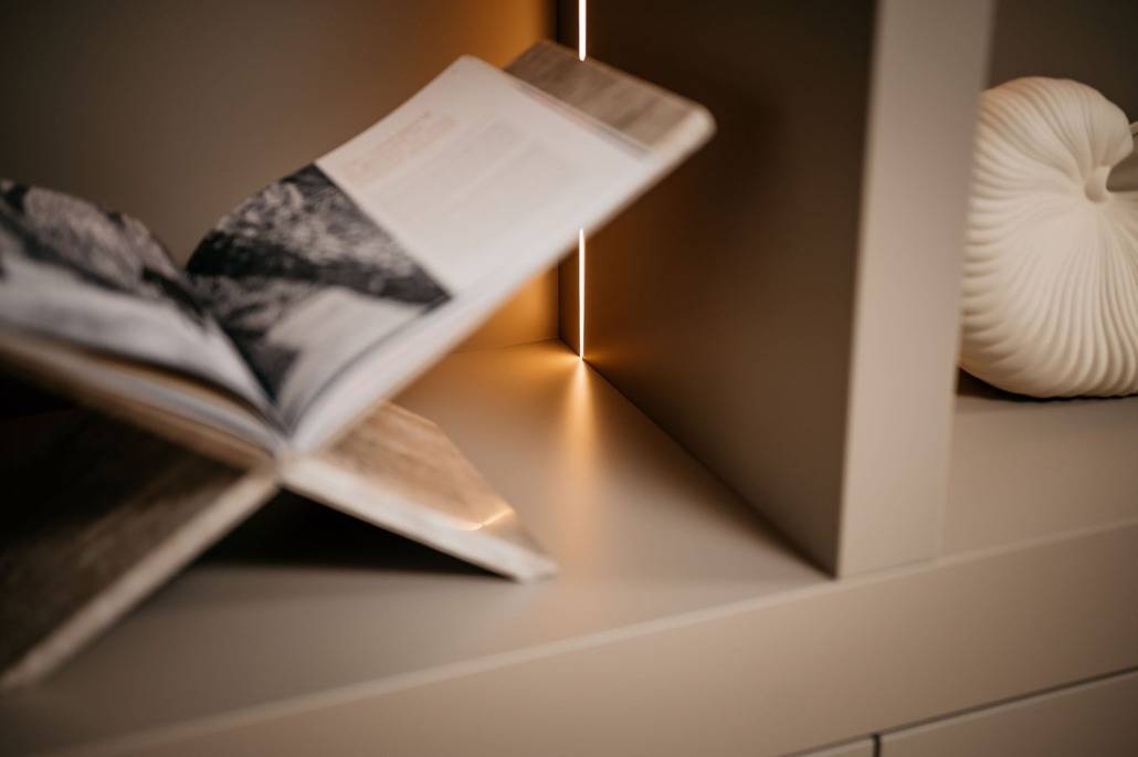 Een open boek rust op een standaard in een beige kast, met zacht warm licht dat uit de hoek schijnt. Een wit decoratief object met structuur staat op de plank rechts van het boek.