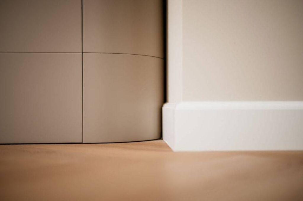 Close-up van een moderne, minimalistische hoek waar een gebogen kast samenkomt met een crèmekleurige muur met een witte plint op een lichte houten vloer.