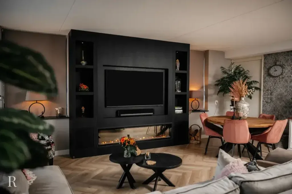 Moderne woonkamer met een grote zwarte entertainmentunit met tv en open haard, ronde eettafel met roze stoelen, lichthouten vloer, planten en neutraal getinte inrichting.