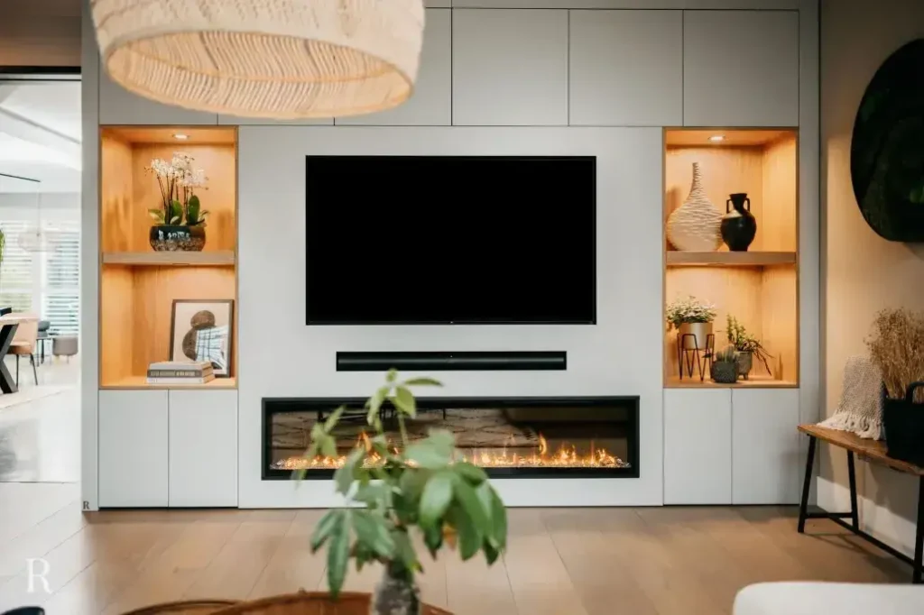 Moderne woonkamer met een ingebouwd wandmeubel met een grote tv, een lineaire open haard eronder en verlichte houten planken met aan weerszijden decoratieve voorwerpen en planten. Een groene potplant staat op de voorgrond.