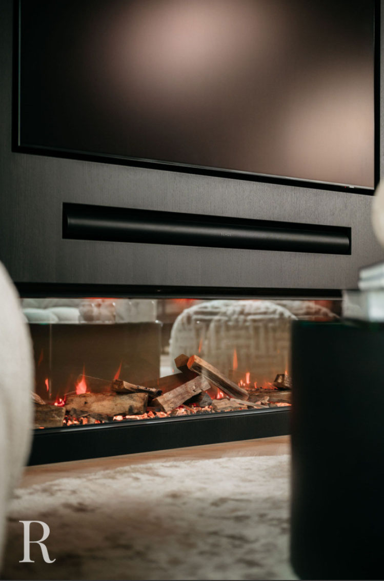 Een moderne elektrische open haard met gloeiende kunstmatige houtblokken en sintels staat onder een strak zwart wandmeubel en een groot tv-scherm in een eigentijdse woonkamer. De letter R is zichtbaar in de linker benedenhoek.