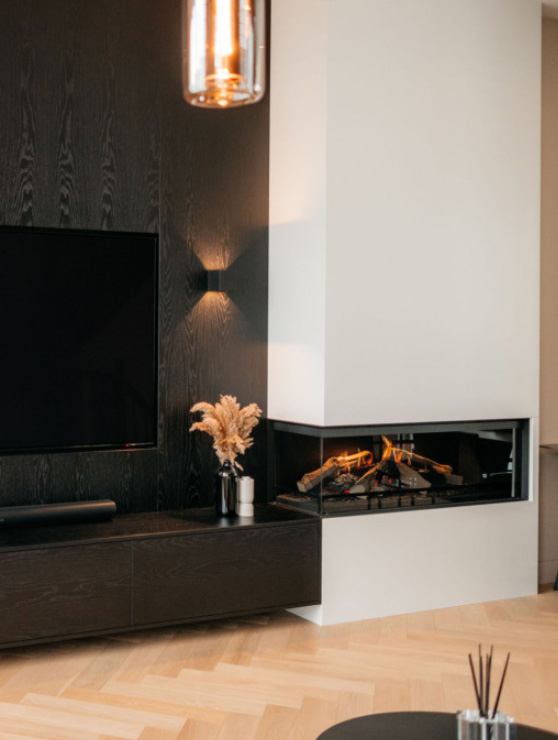 Moderne woonkamerhoek met een tv aan de muur, een strakke zwarte console, een decoratieve vaas met gedroogde planten en een met glas afgesloten open haard in een witte hoekmuur. Daarboven is een hangende lamp zichtbaar.