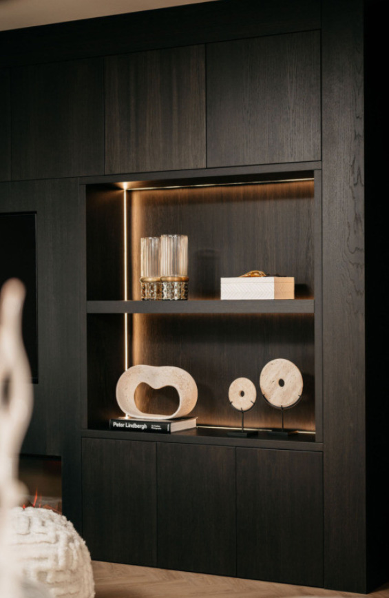 Een modern donkerhouten rek toont decoratieve voorwerpen, waaronder glazen bussen, een doos met textuur, sculpturale stenen stukken en boeken, allemaal zacht verlicht door ingebouwde lampen.