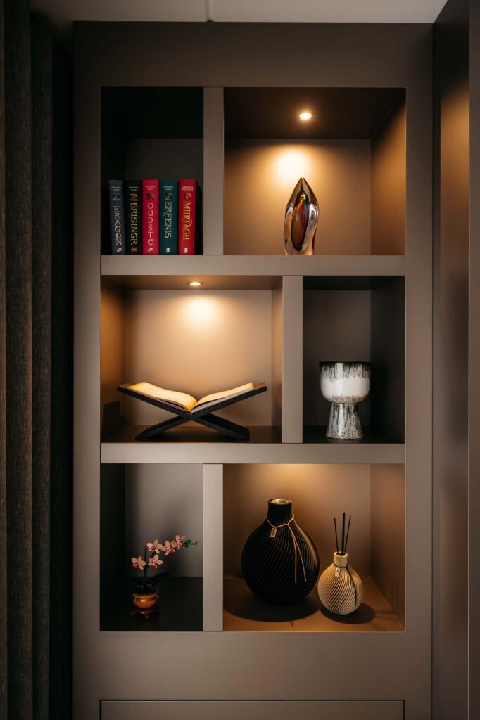 Een moderne Japandi-cinewall bestaat uit een ingebouwde plank met boeken, een open boek, decoratieve vazen, een glazen schaal, een klein bloemstuk en zachte spotjes die de zorgvuldig geselecteerde voorwerpen subtiel verlichten.