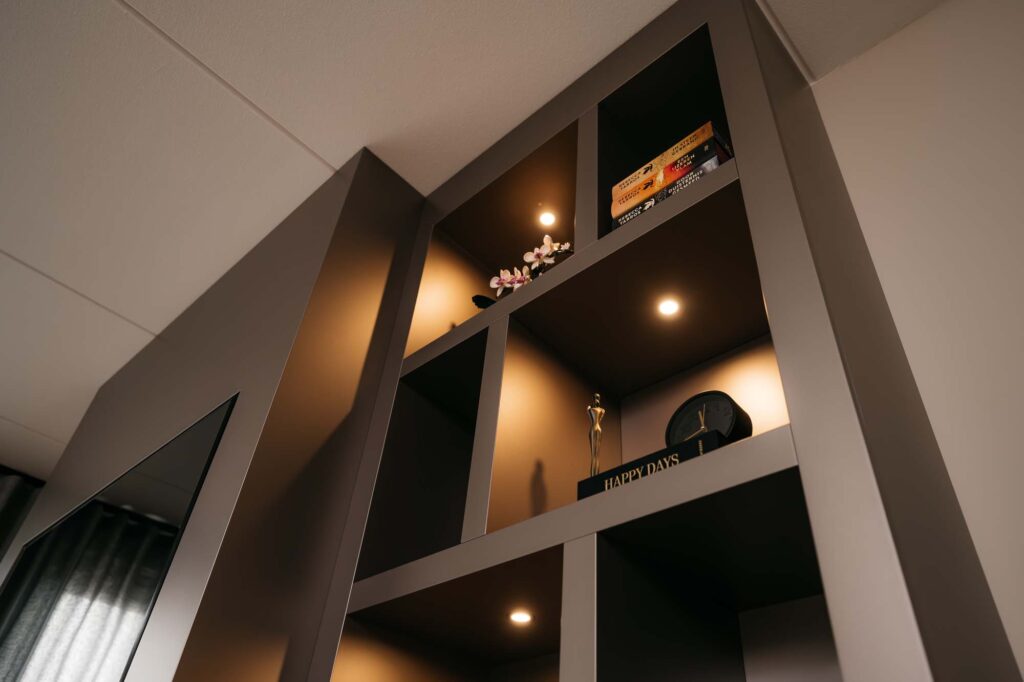 Een moderne, ingebouwde Japandi-boekenkast in cinewall-stijl met spotjes, waarop een klein potplantje, een paar boeken, een klok en een decoratief beeldje staan. De planken hebben een minimalistisch, eigentijds ontwerp.