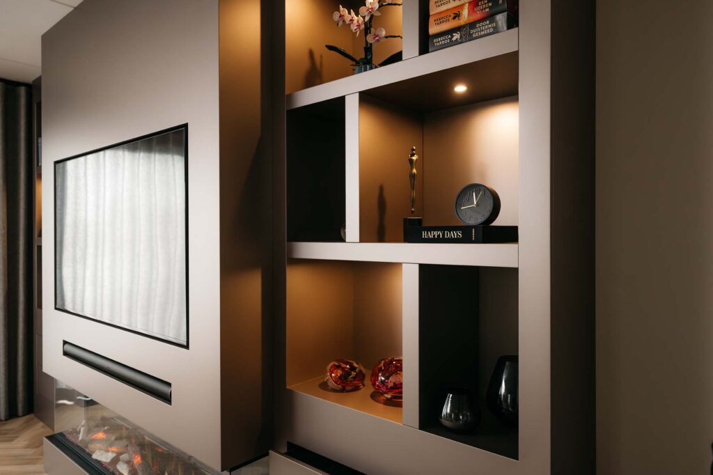 Moderne ingebouwde japandi-boekenkast met decoratieve voorwerpen, waaronder een klok, boeken, kleine beeldjes en een bloemenvaas. Naast de planken bevindt zich een filmwand met daarboven een flatscreen-tv en daaronder een elektrische haard. Warme verlichting zet de inrichting mooi in de schijnwerpers.