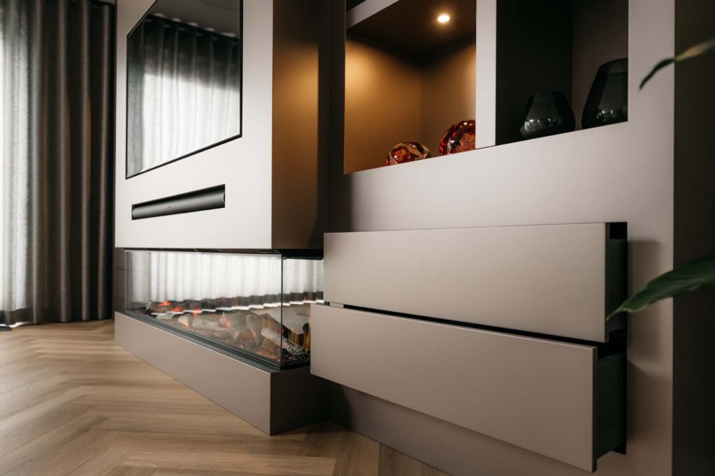 Een moderne woonkamerinrichting met een stijlvolle Japandi-filmwand, voorzien van een ingebouwde elektrische open haard, beige kasten, open planken met decoratieve spullen en daarboven een tv. Een lichte houten visgraatvloer en hoge gordijnen maken het geheel af.