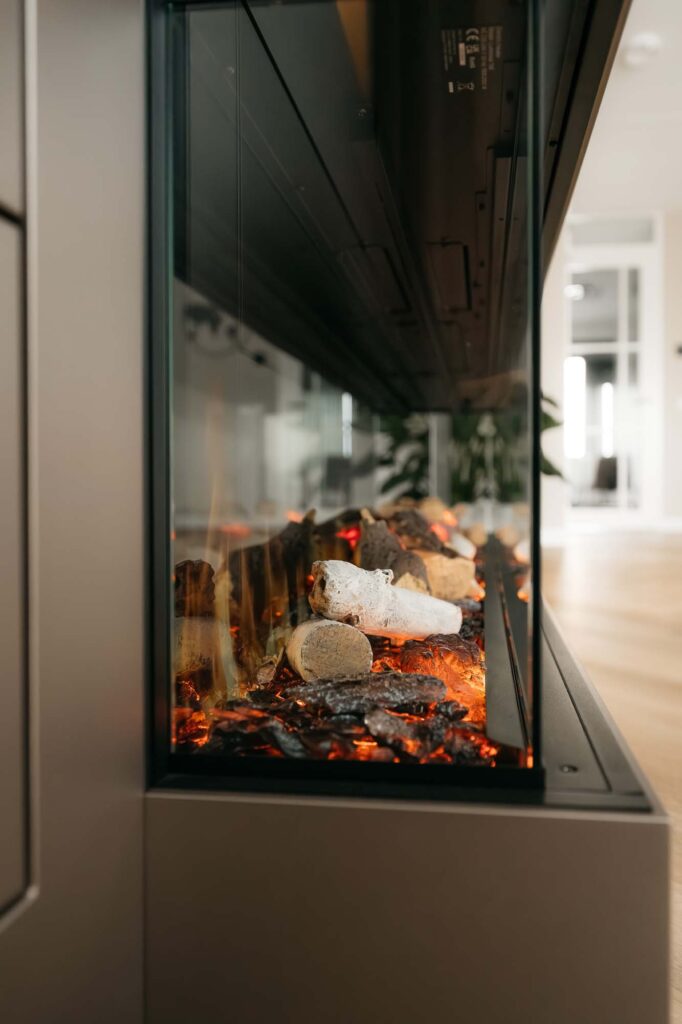Een close-up van de zijkant van een moderne, met glas omgeven open haard met gloeiende sintels en houtblokken, waarbij de Japandi-stijl naadloos is geïntegreerd in een filmwand, wat een warme en gezellige sfeer creëert in een lichte, moderne ruimte.