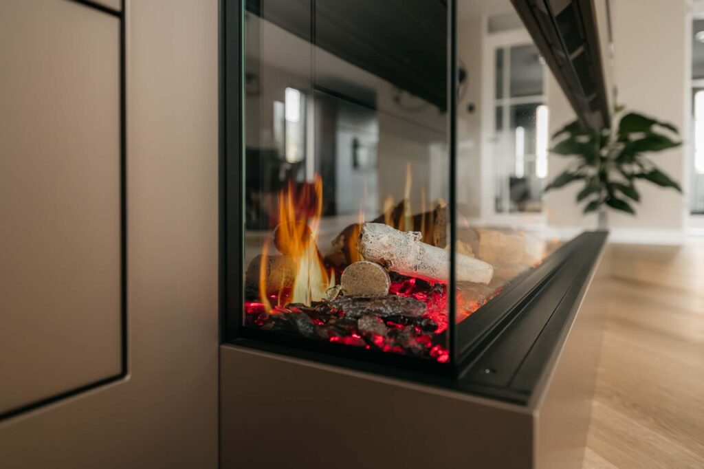 Een moderne, met glas omgeven open haard met realistische vlammen en gloeiende sintels is ingebouwd in een stijlvolle Japandi-filmwand en geeft een eigentijdse woonruimte met houten vloeren en een weelderige plant op de achtergrond extra glans.
