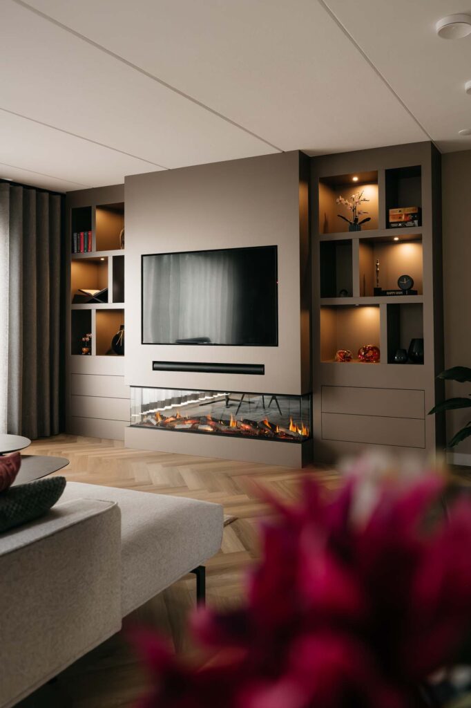 Moderne woonkamer met een op de japandi-stijl geïnspireerde filmwand, voorzien van een grote flatscreen-tv, een elektrische open haard en verlichte planken waarop decoratieve voorwerpen staan uitgestald. Op de voorgrond staat een wazige rode bloem.