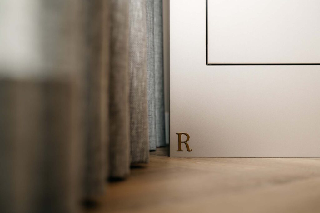 Een close-up van een japandi-filmwand toont een lichtgekleurd paneel met de letter R in goud in de benedenhoek, naast grijze gordijnen die losjes hangen en een houten vloer.