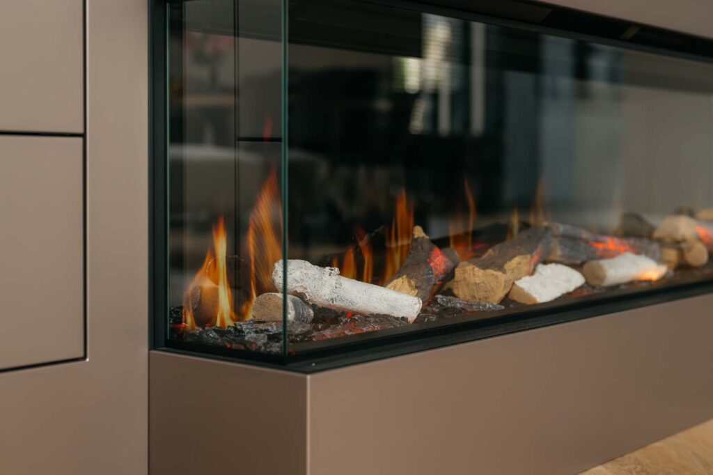 Een moderne, met glas omgeven open haard met realistisch ogende houtblokken en gloeiende vlammen is ingebouwd in een strakke Japandi-filmwand, die perfect past bij de neutrale muren en de overvloed aan natuurlijk licht in deze eigentijdse ruimte.
