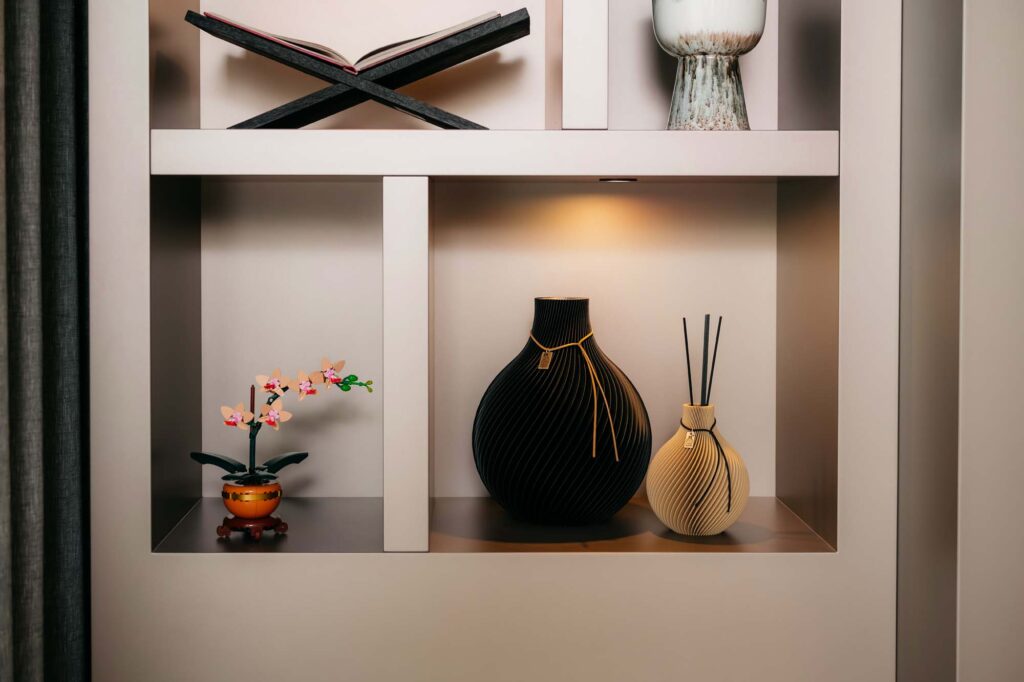 Op een moderne Japandi-plank staan een klein roze kunstbloemetje in een potje, een grote zwarte vaas, een beige vaas met geurstokjes en een open boek op een standaard, allemaal netjes gerangschikt onder warme verlichting naast de elegante cinewall.