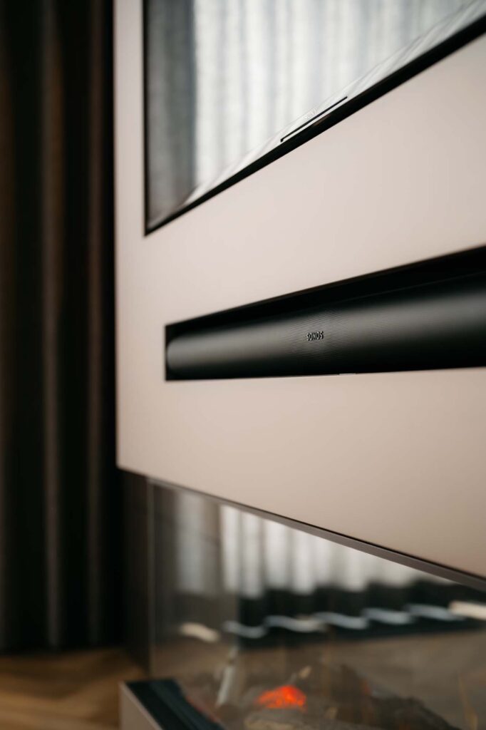 Een close-up van een moderne Japandi-woonkamer met een SONOS-soundbar die onder de televisie is bevestigd aan een strakke cinewall, boven een elektrische open haard, met gordijnen en zonlicht op de achtergrond.