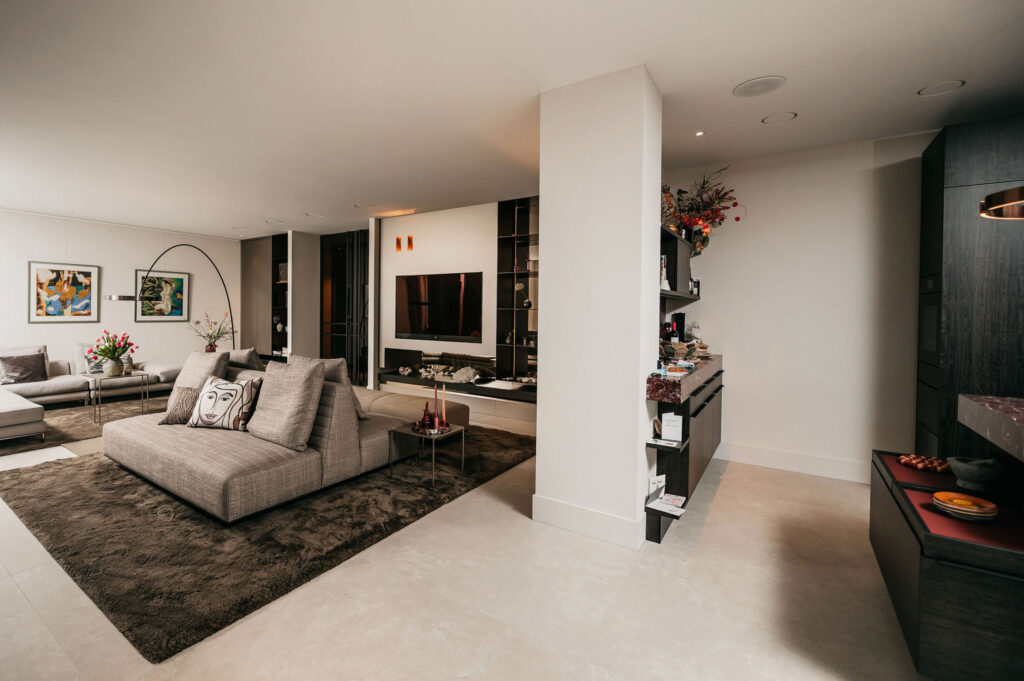 Een moderne, ruime woonkamer met neutrale tinten, een grote grijze bank, bruin vloerkleed, tv aan de muur, ingebouwde planken en eigentijdse inrichting. De kamer is goed verlicht met minimalistisch en stijlvol meubilair.