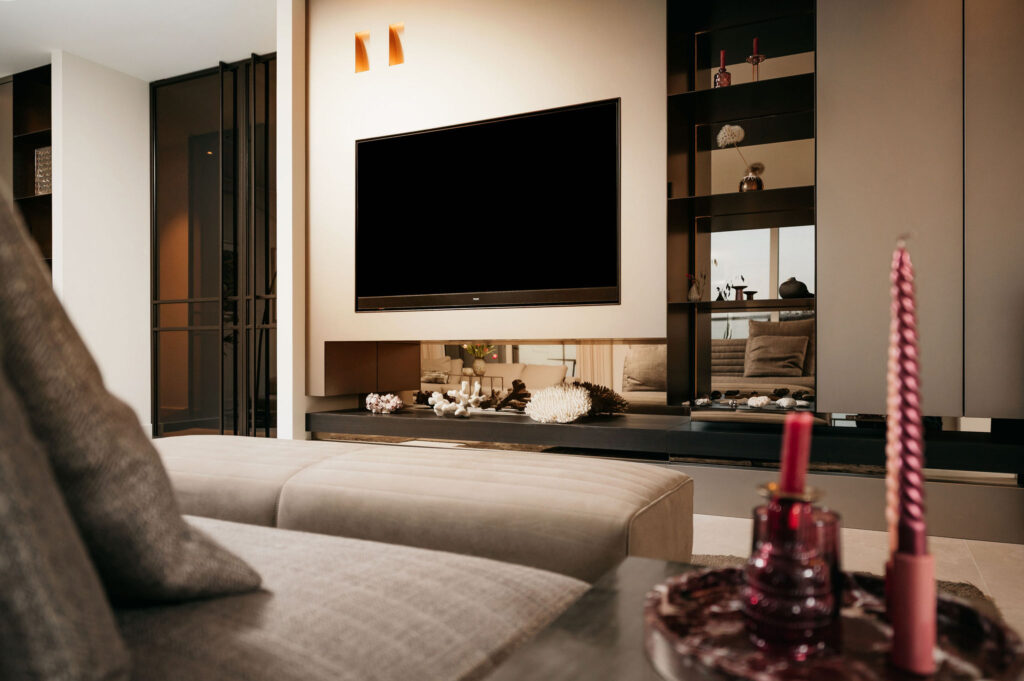 Moderne woonkamer met een aan de muur gemonteerde flatscreentelevisie, decoratieve planken, een beige bank en een salontafel met roze kaarsen en rood glaswerk. Warme verlichting en een nette, hedendaagse inrichting zijn zichtbaar.