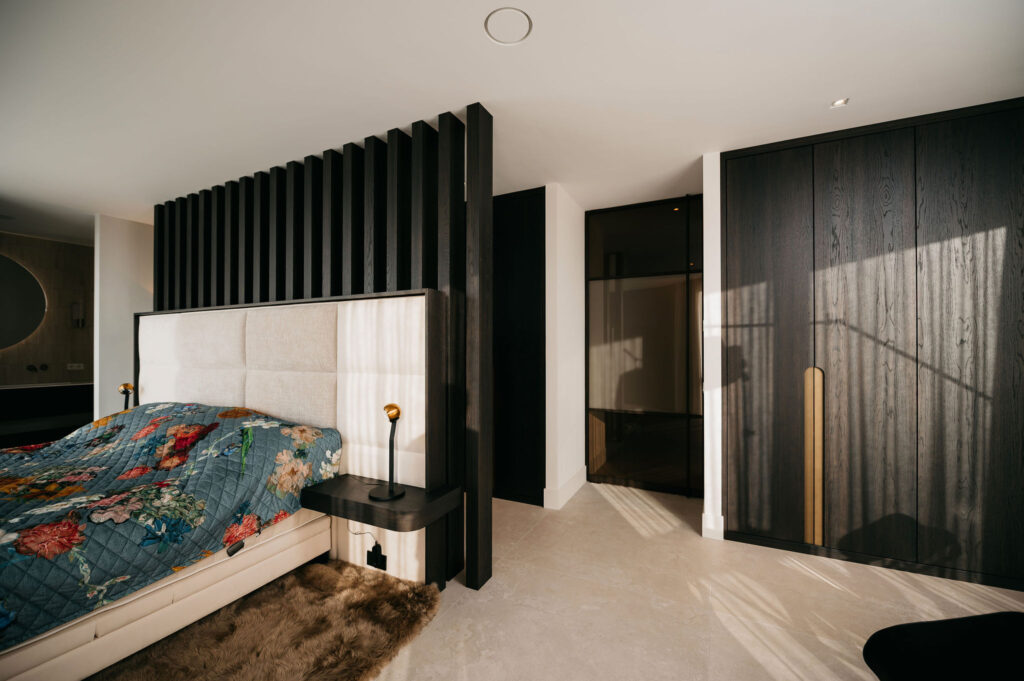 Een moderne slaapkamer met een groot gecapitonneerd hoofdeinde, donkere verticale latten, een gebloemde sprei, gouden bedlampjes, een bontkleed en zonlicht dat op bleke vloeren en zwarte houten deuren valt.