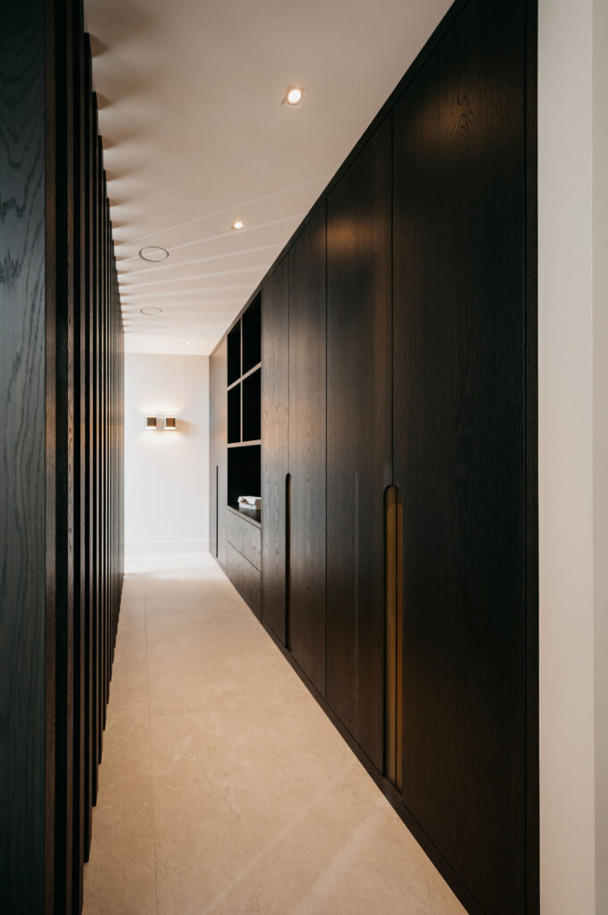Een moderne hal met lichtbeige vloeren, donkere houten inbouwkasten rechts en verticale houten latten links. Inbouwverlichting en een wandarmatuur zorgen voor verlichting in de minimalistische ruimte.