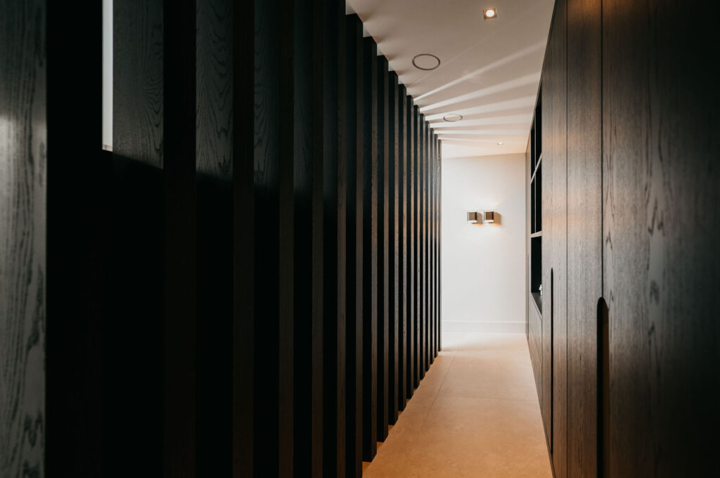 Een moderne hal met donkere verticale houten latten aan de linkerkant en gladde donkere muren aan de rechterkant, die leidt naar een goed verlichte witte muur met een eigentijdse wandlamp.