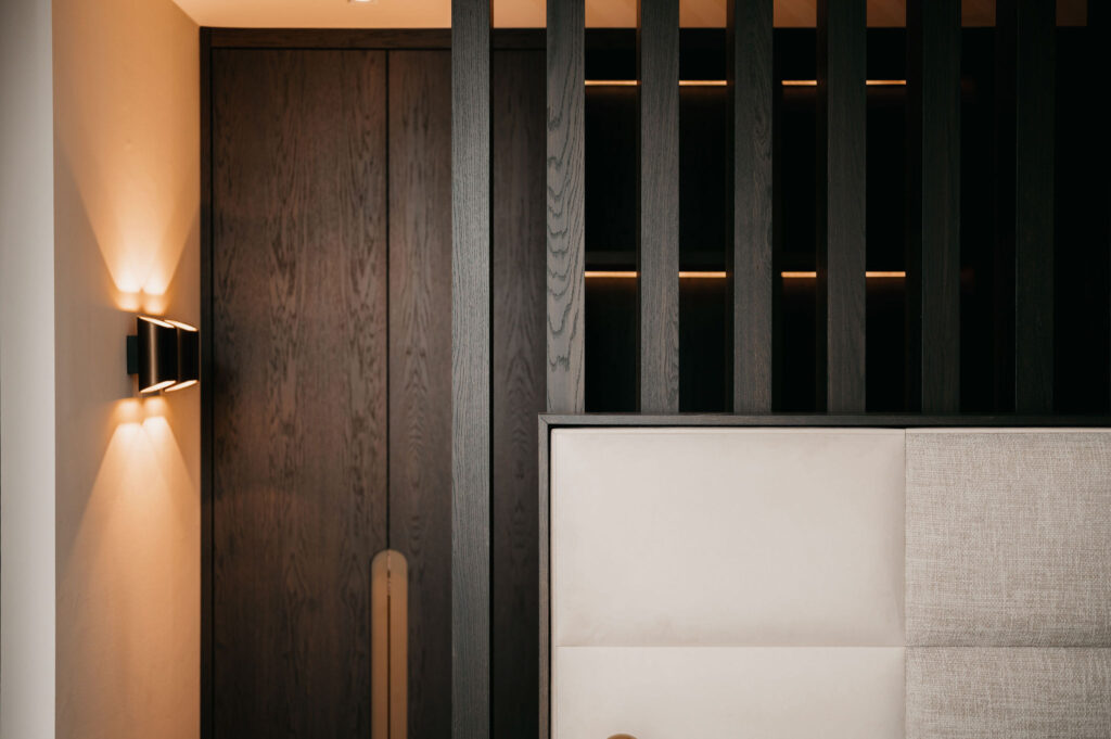 Een modern interieur met verticale donkerhouten latten, een zwarte houten deur, zachte ingebouwde verlichting en een deel van een lichtbeige gestoffeerd hoofdeinde op de voorgrond.