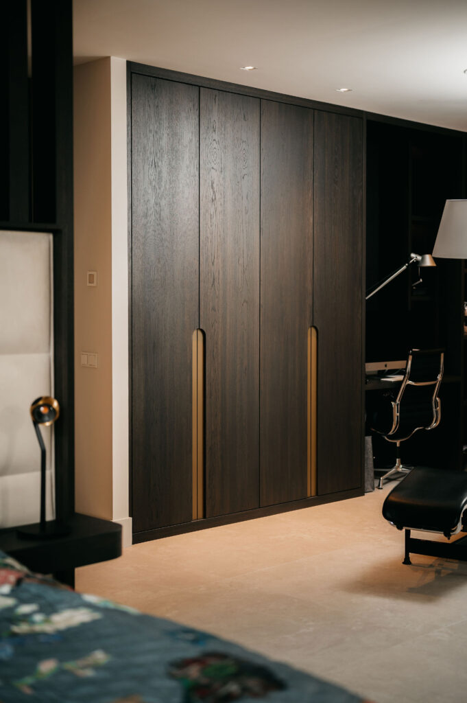 Een moderne, donkerhouten kledingkast met hoge verticale gouden handgrepen staat tegen een beige muur in een hedendaagse slaapkamer, met een deel van een bed en een zwarte stoel zichtbaar op de voorgrond.