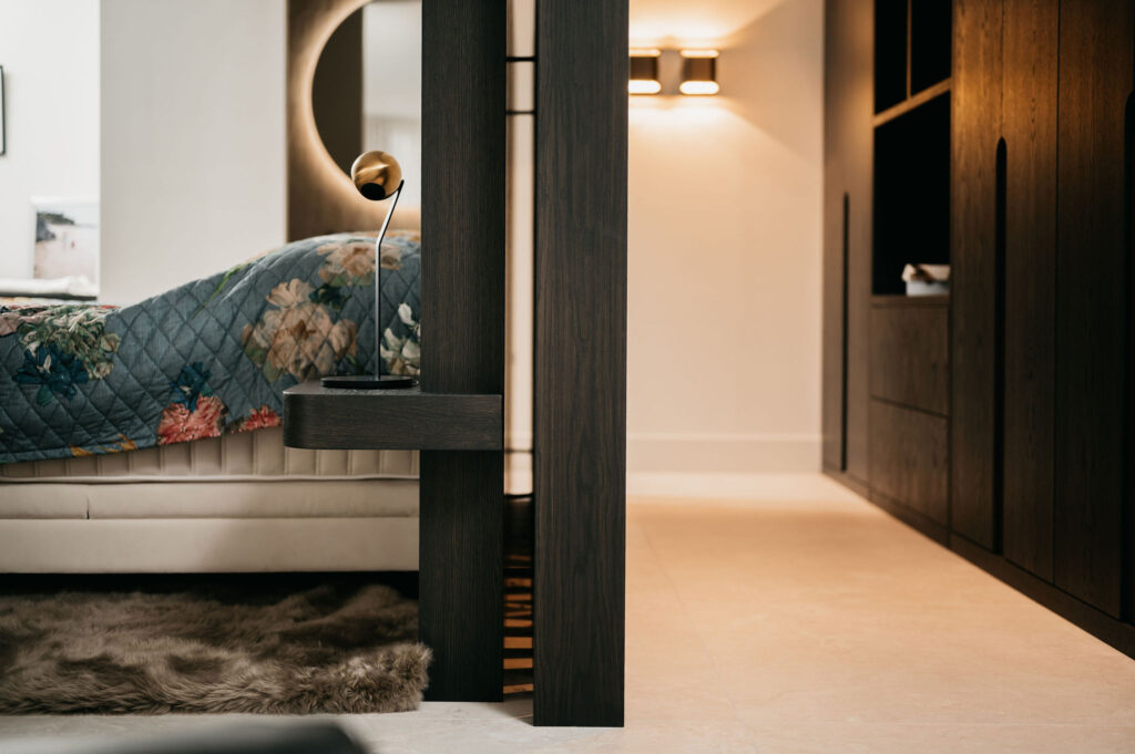 Moderne slaapkamer met een scheidingswand en ingebouwde plank, een bed met een gebloemd dekbed, een bontkleed en warme verlichting. De inrichting is eigentijds met neutrale tinten en strakke lijnen.