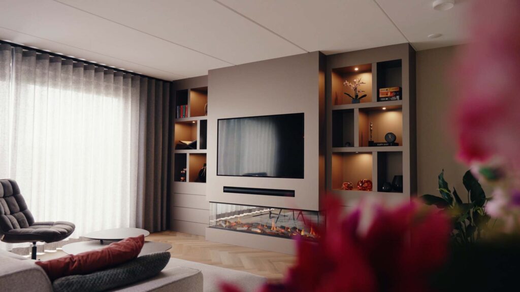 Een moderne woonkamer met een gezellige grijze bank, een op de japandi-stijl geïnspireerde filmmuur met een aan de muur bevestigde flatscreen-tv boven een strakke open haard, ingebouwde planken met decoratieve voorwerpen en grote ramen met transparante gordijnen die natuurlijk licht binnenlaten.