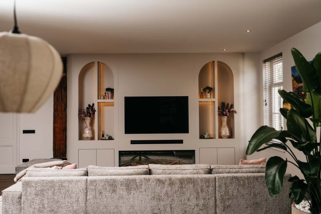 Moderne woonkamer met een grote grijze hoekbank, een aan de muur bevestigde tv, ingebouwde planken met vazen en planten, sfeervolle verlichting, een grote groene plant en een raam met jaloezieën dat natuurlijk licht binnenlaat.