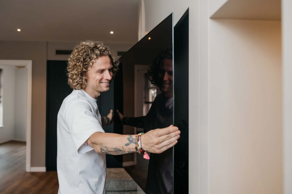Een glimlachende persoon met krullend haar en tatoeages hangt een flatscreen-tv aan de muur in een moderne woonkamer.