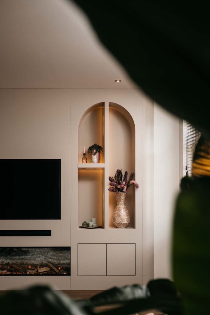 Een muur in de woonkamer met een zwarte televisie en een lineaire open haard. Daarnaast bevindt zich een ingebouwde, crèmekleurige nis met boogvormige opening, voorzien van warme inbouwverlichting, een mozaïekvaas met gedroogde bloemen, kaarsen en een klein sierplantje.