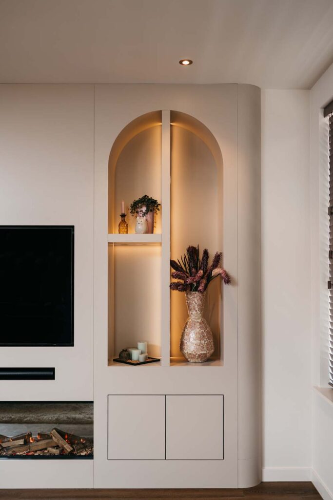 In een ingebouwde wandnis met sfeervolle verlichting staan decoratieve vazen, gedroogde bloemen en kaarsen tentoongesteld. De nis, die grenst aan een aan de muur bevestigde tv en een moderne open haard, geeft de eigentijdse woonkamer een elegante uitstraling.