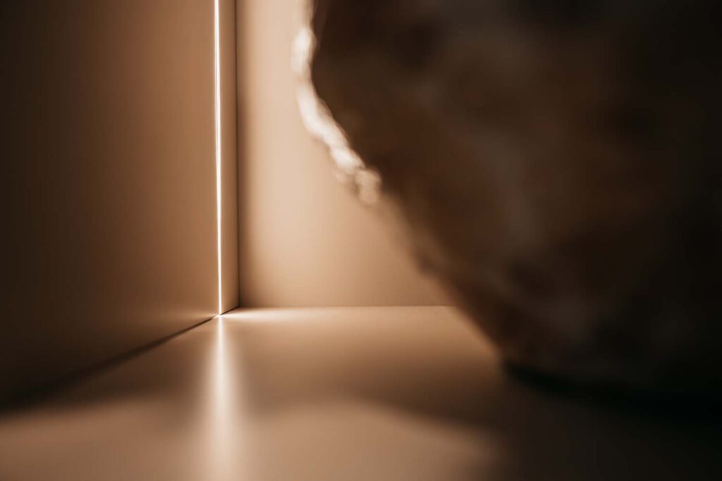 Zacht licht schijnt door een smalle opening tussen twee beige muren en weerkaatst op een glad oppervlak. Aan de rechterkant van de afbeelding is een wazig, rond voorwerp te zien.