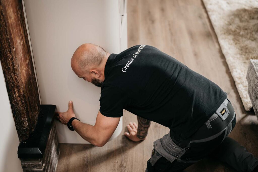 Een man in een zwart shirt en een werkbroek zit op zijn knieën op een houten vloer en is bezig met het plaatsen of controleren van een witte kast bij een open haard.