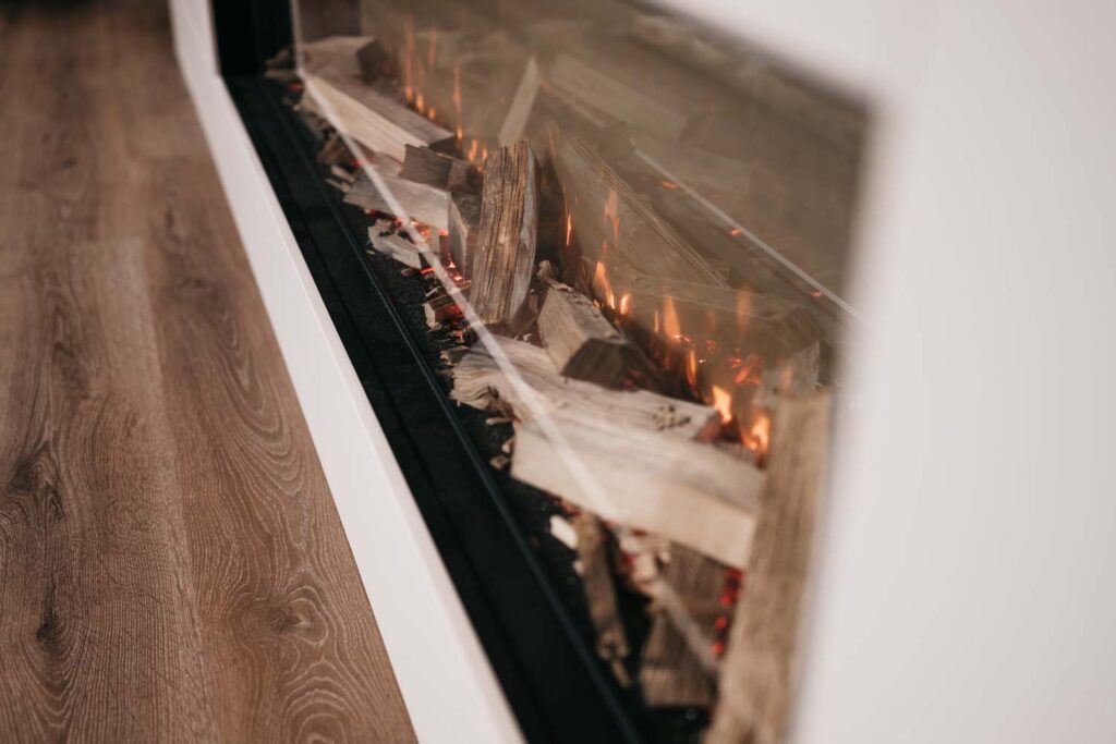 Moderne open haard met een glazen voorkant, brandende houtblokken en kleine oranje vlammen. De open haard is ingebouwd in een witte muur, met links een houten vloer.