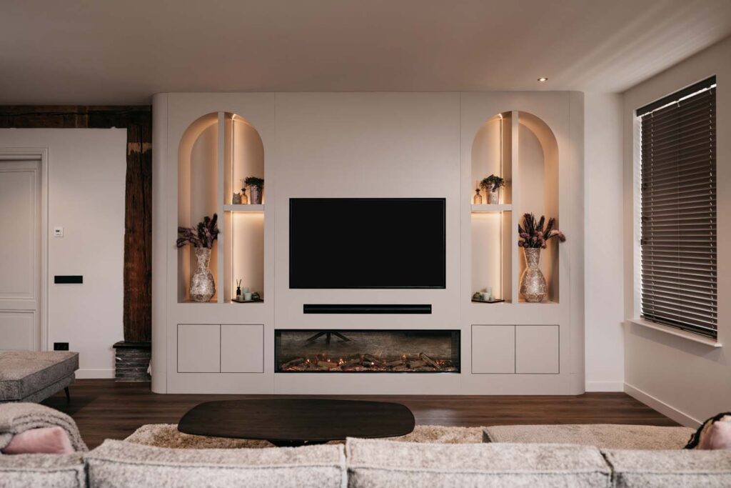 Moderne woonkamer met een aan de muur bevestigde tv, ingebouwde planken met vazen en planten, daaronder een elektrische open haard, neutrale banken, een donkere salontafel en grote jaloezieën die natuurlijk licht binnenlaten.