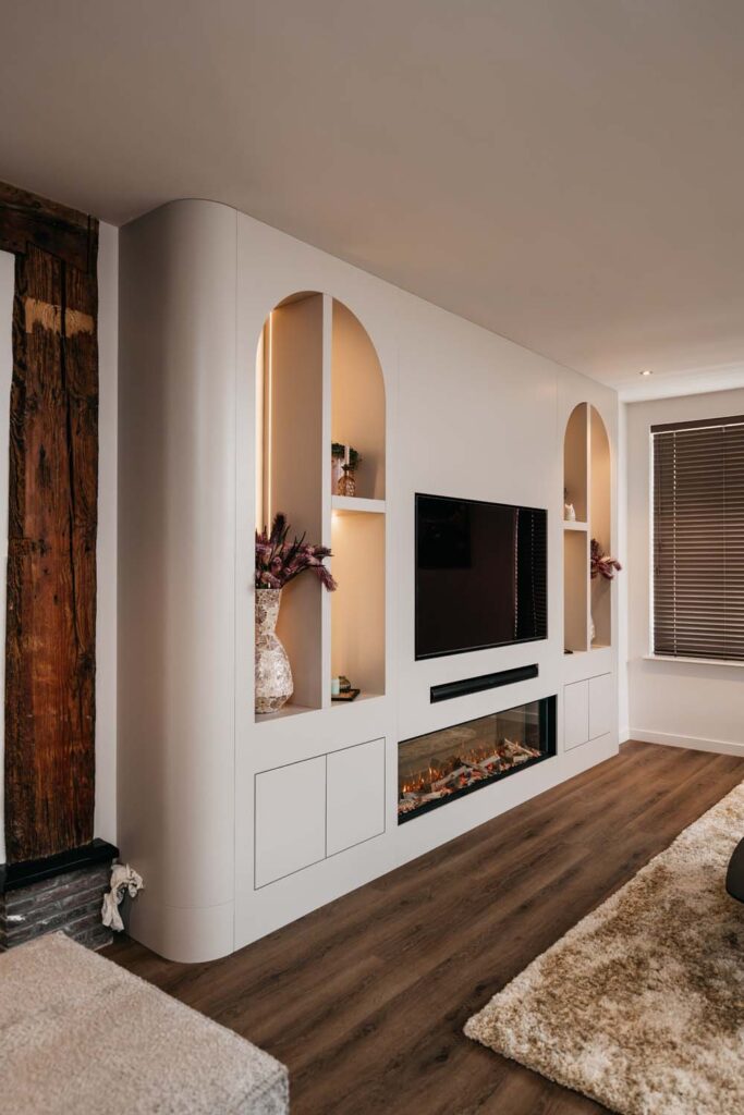 Moderne woonkamer met een ingebouwd wit tv-meubel met daarin een tv, een open haard, planken met vazen en bloemen, en een houten vloer. Ook zijn er een hoogpolig tapijt en een houten wandaccent te zien.
